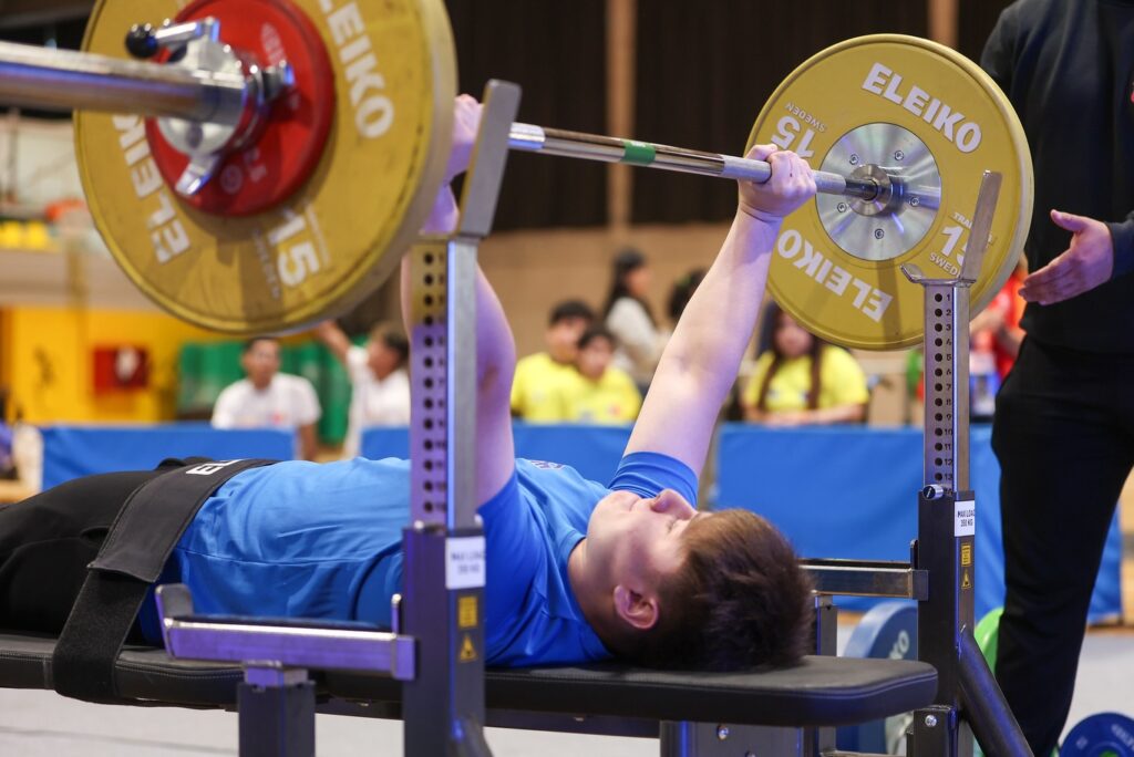 Fotografía de joven con polera azul acostado realizando un levantamiento en para powerlifting. 